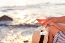 Girl is sitting on sea beach with wineglass of wine at sunset in summer vacation in resort. Tourist woman in striped dress with straw hat is enjoying life, relaxing, drinking, traveling.